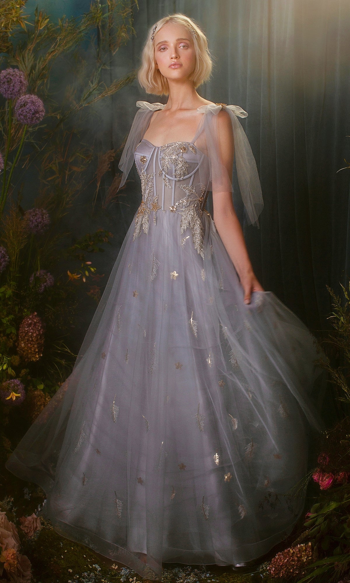 A woman, surrounded by tall flowers and greenery in soft lighting, wears the Shoulder-Bow Long Corset Prom Ball Gown A0824 with a light blue tulle skirt, floral embroidery, corset bodice, and sheer shoulder ribbons.